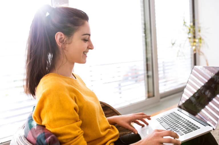 A woman in a yellow sweater smiling while typing content on a laptop.