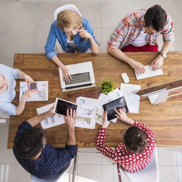 Five people sitting around a wooden table, using laptops and tablets, viewed from above. Office environment with papers and potted plant on the table.